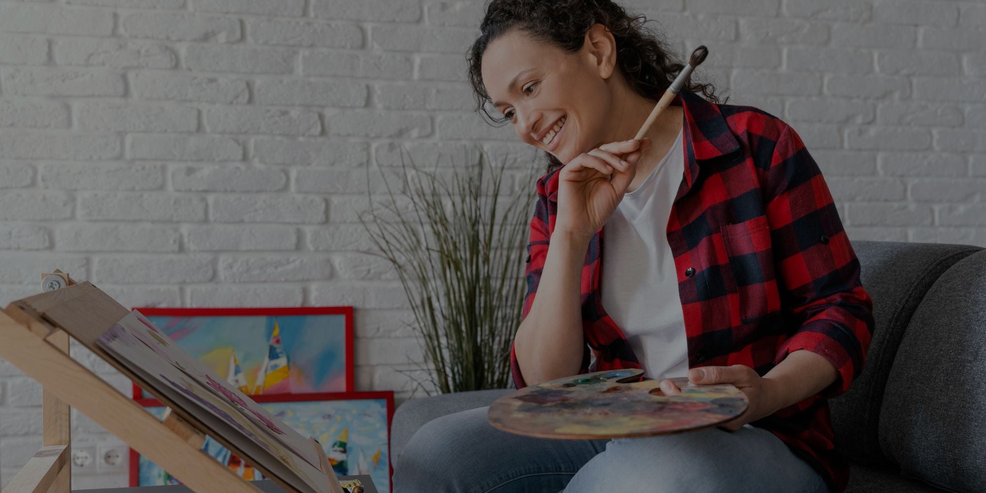 Woman sitting on a couch holding a paintbrush and palette, with art supplies and a plant in the background.